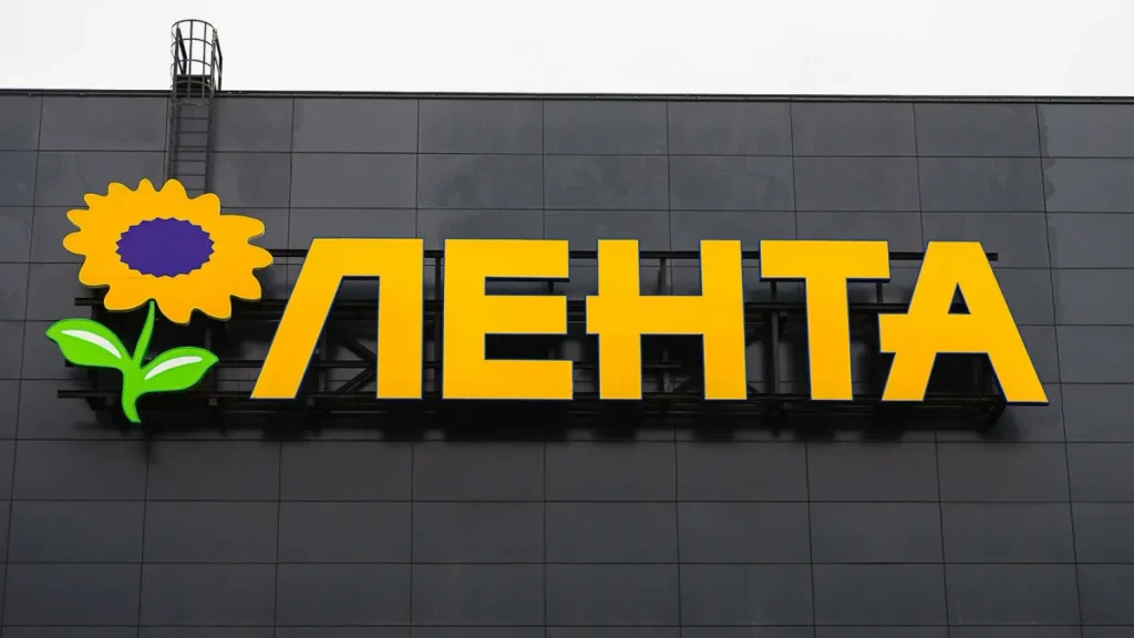 Вывеска «Лента» с жёлтыми буквами и цветком на сером фасаде